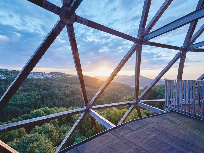 Sonnuntergang sichtbar durch ein geometrisches Stahlgitter mit Wald und Hügeln im Hintergrund.