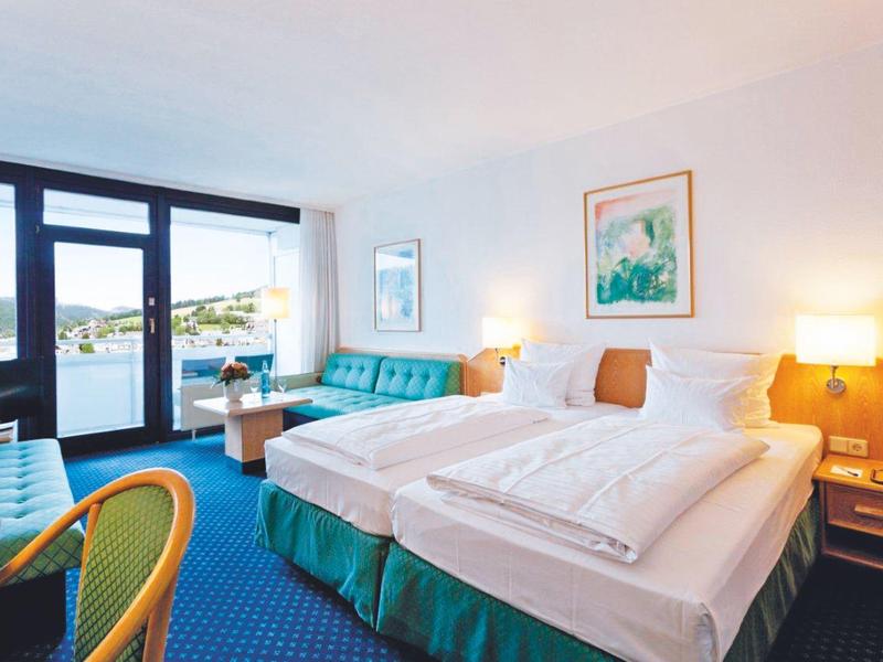 Hell eingerichtetes Hotelzimmer mit zwei Einzelbetten, blauen Sitzmöbeln und großem Fenster.