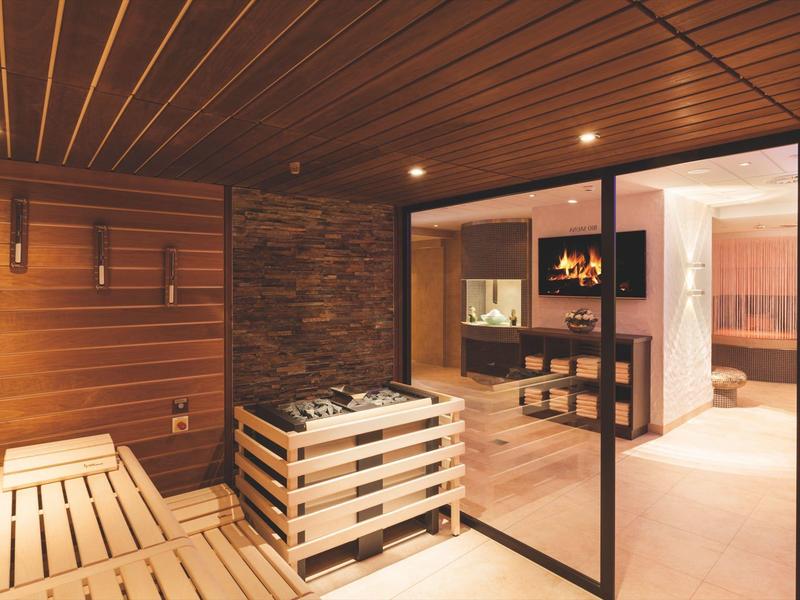 Moderne Sauna mit Holzverkleidung, Steinofen und angrenzendem Ruheraum mit Kaminfeuer.