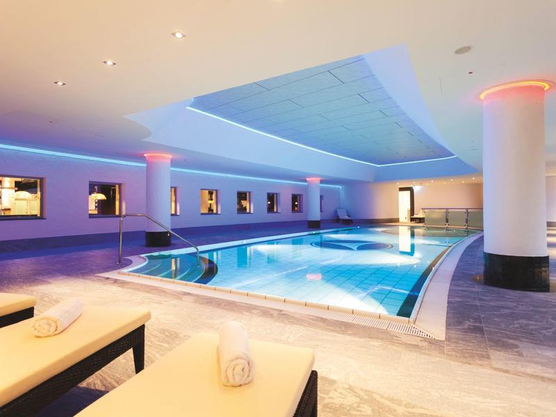 Moderner Innenpool mit blauer Beleuchtung, Liegen und weißen Säulen in hellem Ambiente.