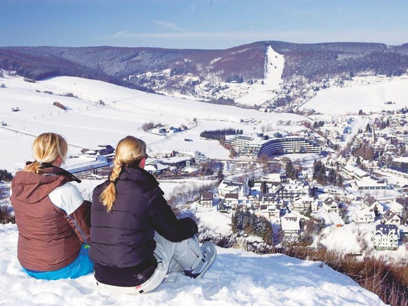 Zwei Personen sitzen im Schnee und blicken auf ein verschneites Tal mit Häusern und Bergen.