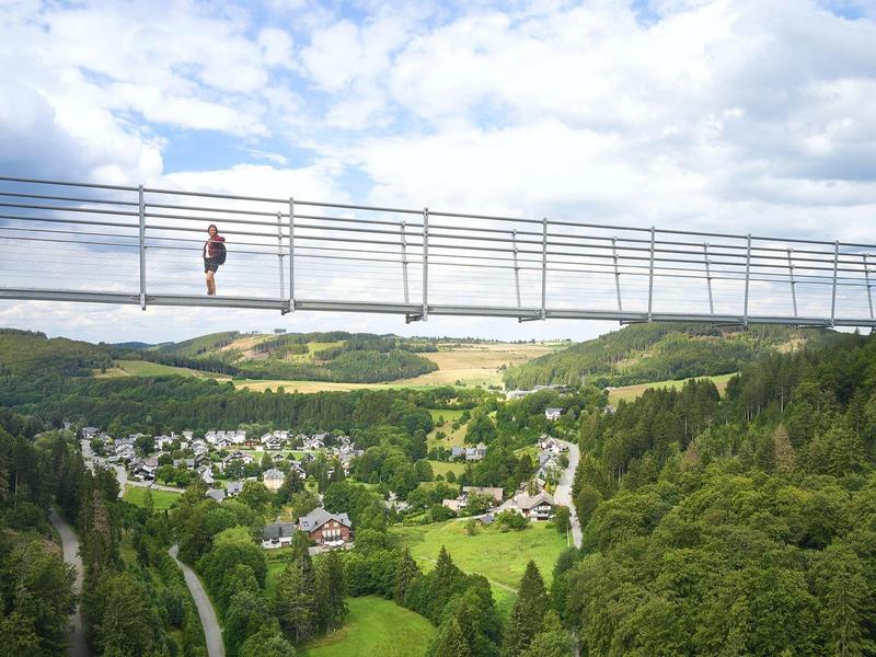 Hängebrücke über grünes Tal mit Wald, Dorf, Hügeln und teils bewölktem Himmel im Hintergrund.
