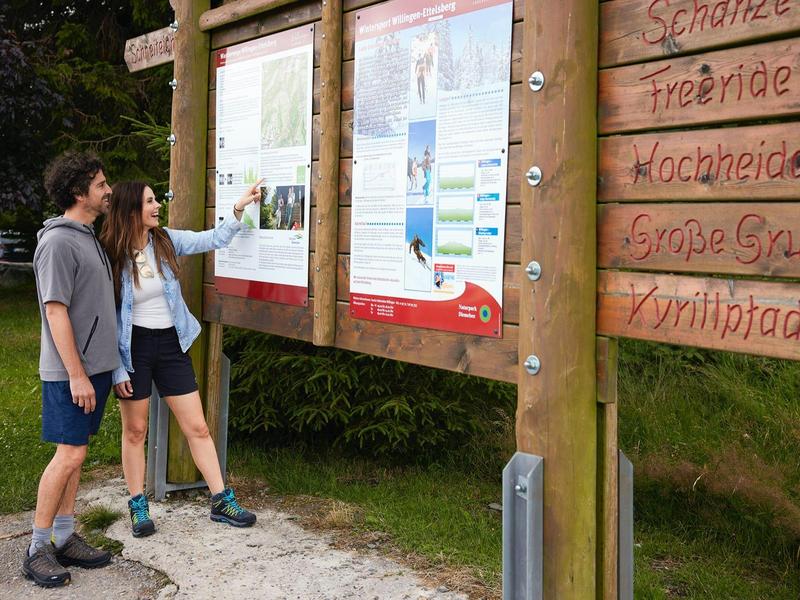 Zwei Wanderer lesen Karten und Informationen an einem großen hölzernen Infostand im Wald.