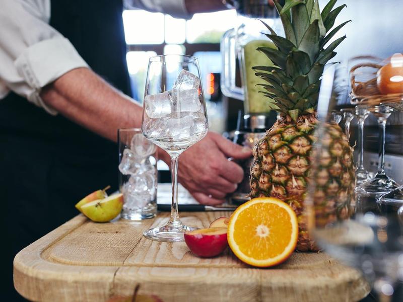 Eine Ananas, eine halbe Orange, ein Cocktailglas mit Eis und eine Person am Holzbrett.