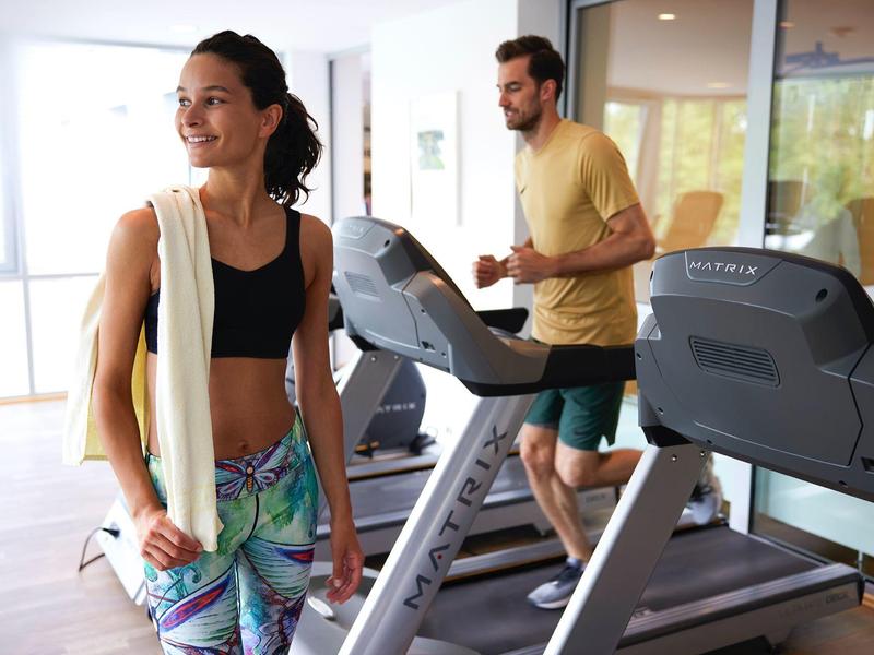 Eine Frau mit Yogamatte geht lächelnd vorbei, während ein Mann auf einem Laufband trainiert.
