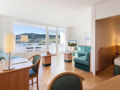 Lichte hotelkamer met bureau, zithoek en balkon met uitzicht op de bergen.