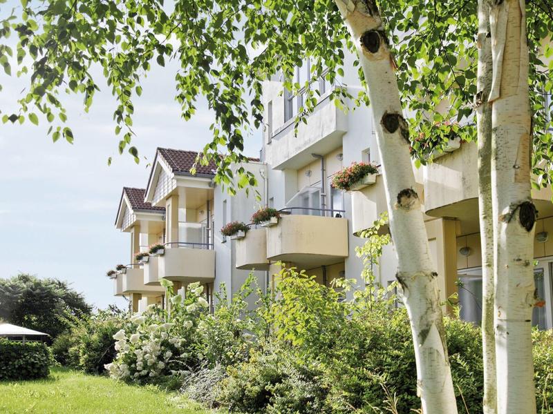 Mehrfamilienhaus mit Balkonen, umgeben von grünen Büschen und zwei Birken im Vordergrund.