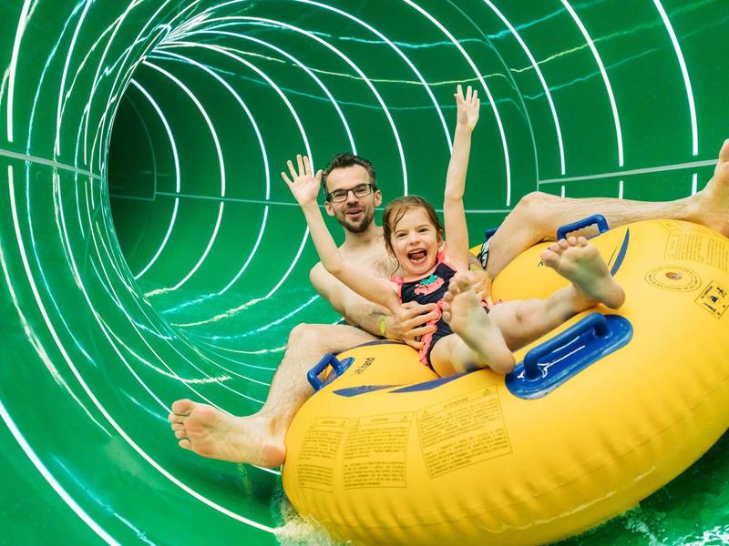 Zwei Personen fahren lachend auf einer gelben Reifenröhre durch eine grüne Wasserrutsche.