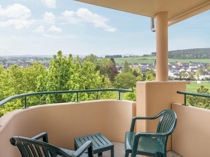 Balkon mit grünem Geländer, zwei grünen Stühlen und Tisch, grüner Landschaft und Häuser im Hintergrund.