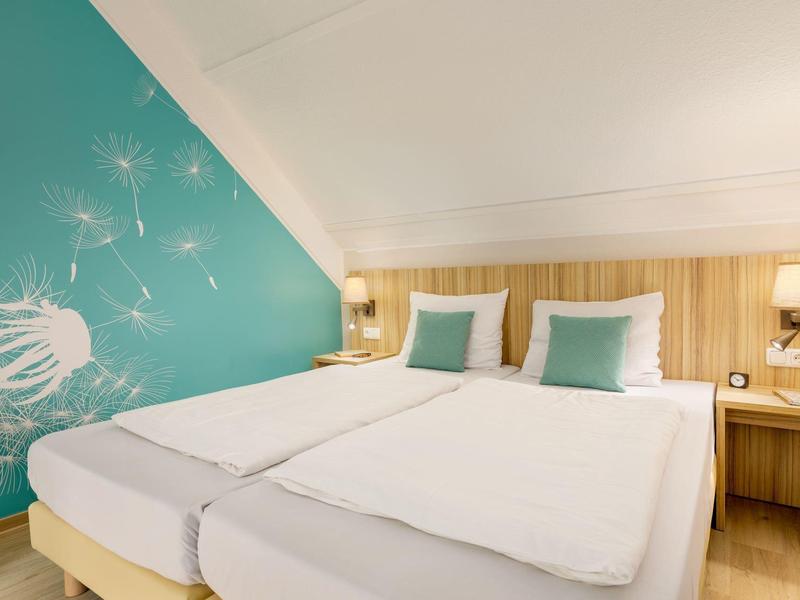 Modernes Schlafzimmer mit zwei Einzelbetten, hellen Holzmöbeln und türkisfarbener Wand mit Pusteblumenmotiv.