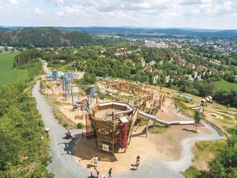 Abenteuer-Spielplatz mit Seilgärten, Türmen und Kletterelementen umgeben von Wald und Stadtlandschaft.