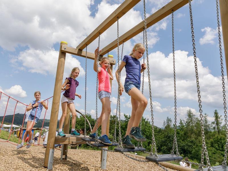 Kinder balancieren auf Holzträgern, die an Ketten hängen, auf einem Spielplatz im Freien.