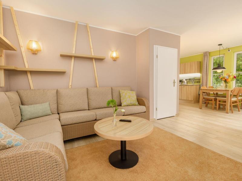 Gemütliches Wohnzimmer mit beige Ecksofa, rundem Holztisch und offenen Regalen an heller Wand.