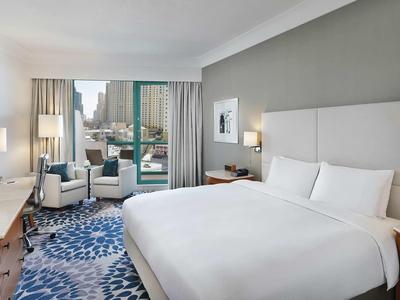 Helles Hotelzimmer mit großem Bett, blauer Teppich, zwei Sesseln und Blick auf Stadt.