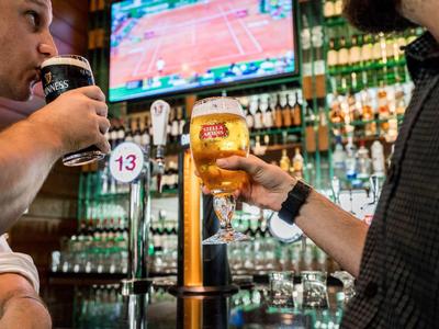 Zwei Männer bestellen Bier an einer Bar mit einem Bildschirm im Hintergrund.