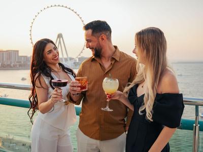 Drei Personen stoßen mit Cocktails auf einer Terrasse mit Meerblick und einem Riesenrad im Hintergrund an.