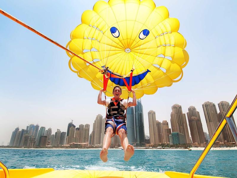Person mit gelbem Fallschirm, der lächelnd aussieht, gleitet über blaues Wasser vor Wolkenkratzern.