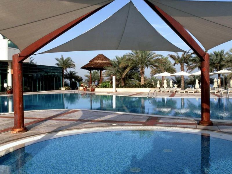 Ein heller Außenpool mit blauen Fliesen, schattenspendenden Segeln und Palmen im Hintergrund.