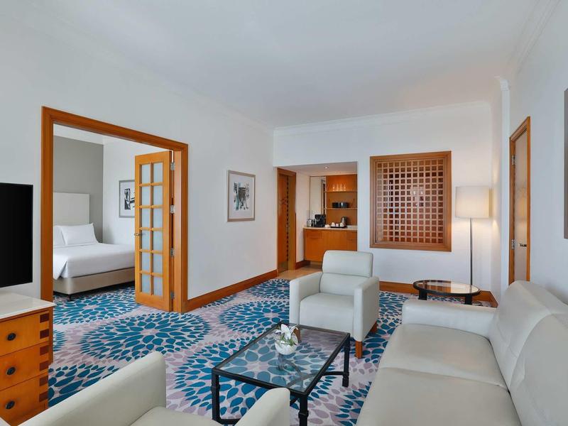 Hell eingerichtetes Wohnzimmer mit blauem Teppich, hellen Sofas, Glastisch und angrenzendem Schlafzimmer.