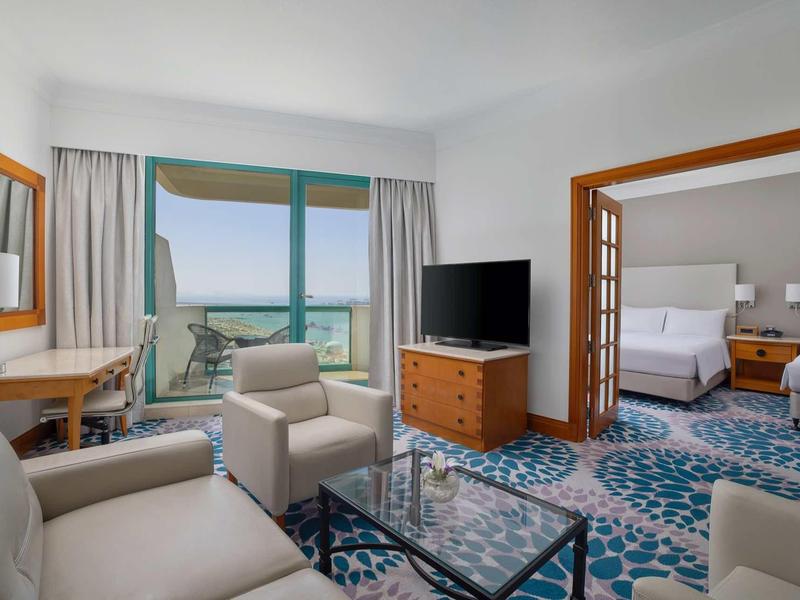 Helles Wohnzimmer mit blau gemustertem Teppich, beige Möbeln, Glas-Couchtisch und TV, angrenzend an Schlafzimmer.