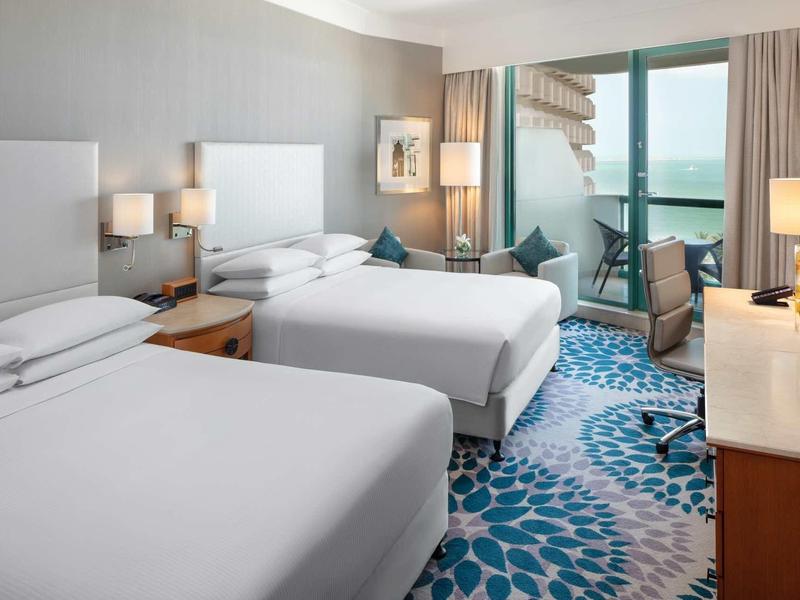 Hell eingerichtetes Hotelzimmer mit zwei Doppelbetten, blauem Teppich und Balkon mit Meerblick.