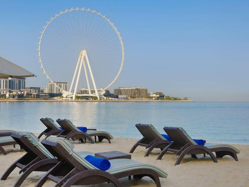 Strand mit Liegestühlen und blauen Handtüchern, im Hintergrund großes Riesenrad und klare blaue See.