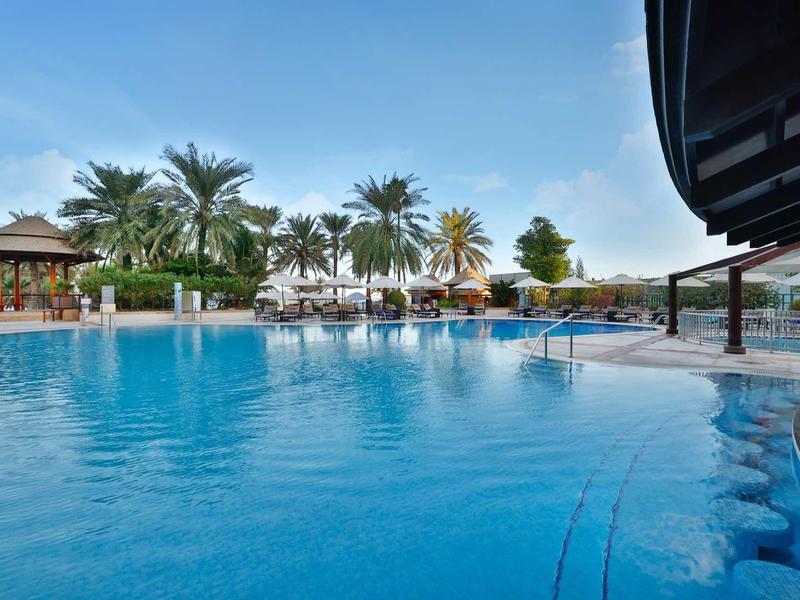 Großer blauer Pool mit Palmen, Himmel und Poolbar in tropischer Hotelanlage bei klarem Wetter.