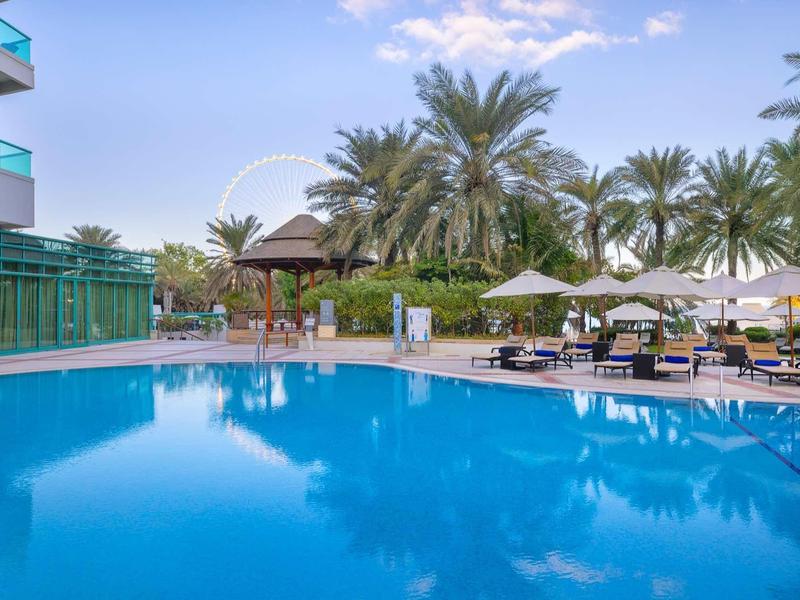 Großer blauer Pool mit Sonnenliegen, weißen Sonnenschirmen und Palmen an bewölktem Himmel.