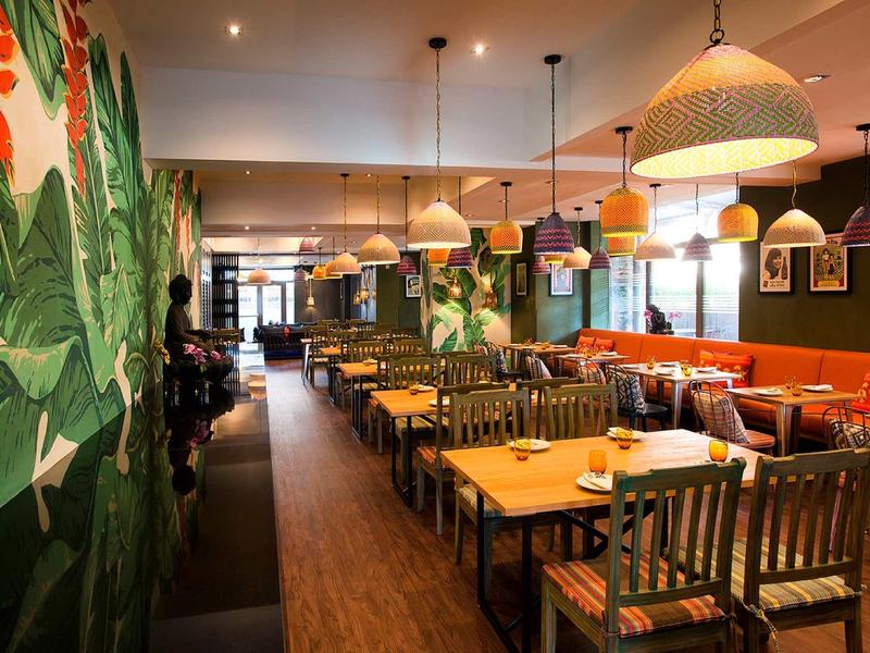 Gemütliches Restaurant mit Holzstühlen, bunten Wänden und großen Pflanzendekoren an der Wand.