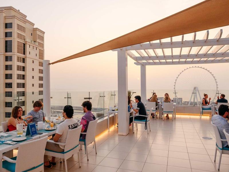 Dachterrasse mit Gästen, modernen Möbeln, offenem Blick auf Stadt und Ferris Wheel im Abendlicht.