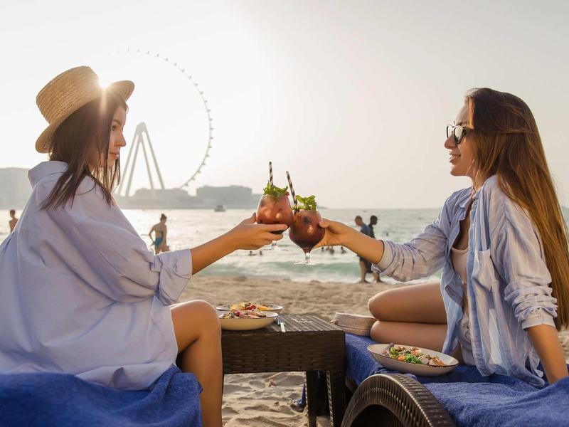 Zwei Frauen sitzen am Strand, stoßen mit Getränken an, Sonne und Riesenrad im Hintergrund.