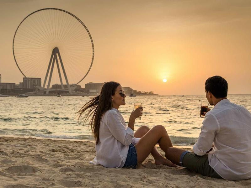 Paar sitzt am Strand bei Sonnenuntergang, im Hintergrund großes Riesenrad am Wasser.