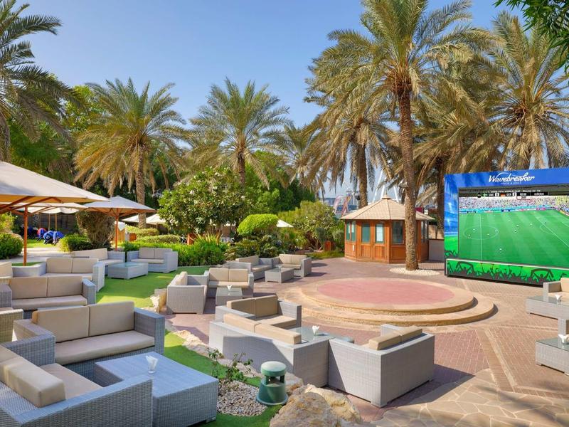 Outdoor-Lounge mit grauen Sofas, Palmen und großer Leinwand bei strahlendem Sonnenschein.