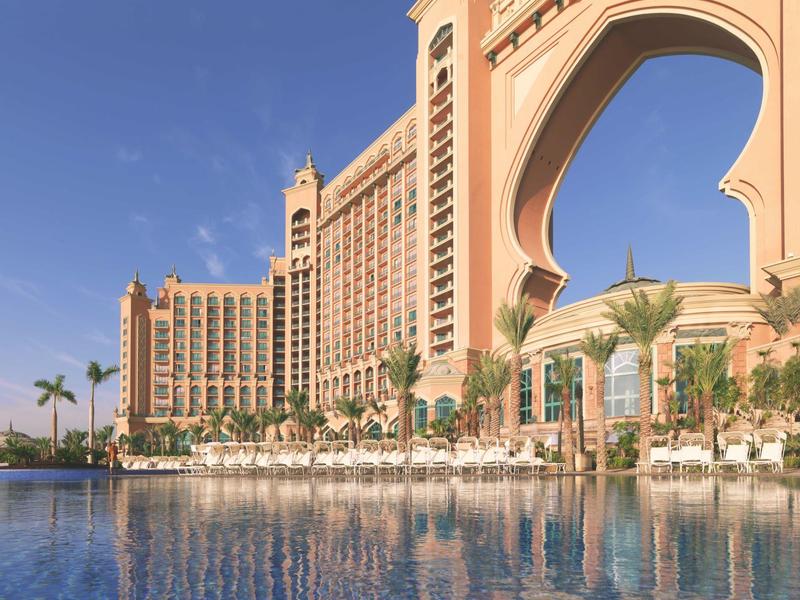 Luxuriöses Hotel mit Palmen und großem Pool, blauer Himmel, helle Fassade, Bögen und Türme.