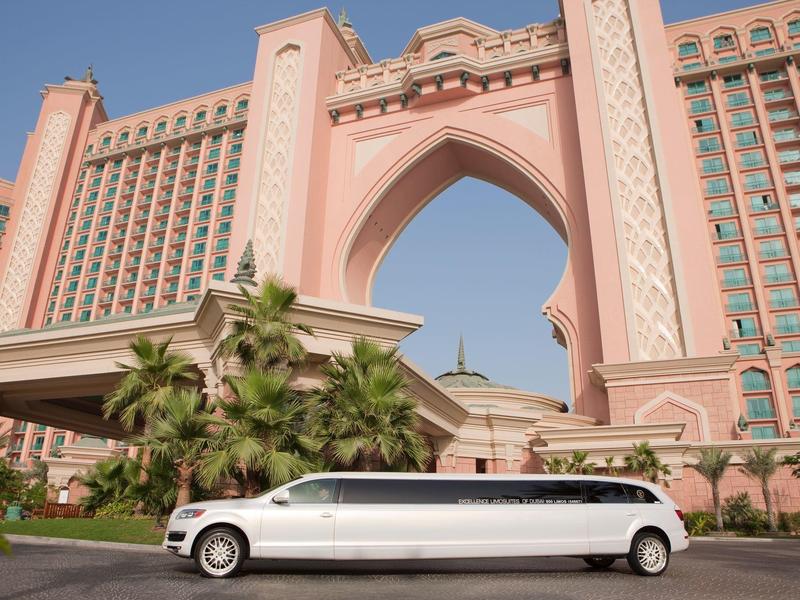 Langer weißer Limousinenwagen vor rosafarbenem Hotel mit großem Bogen und Palmen.