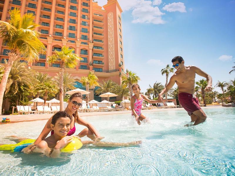 Familie spielt im Pool eines Hotels bei sonnigem Himmel mit Palmen und Liegestühlen.