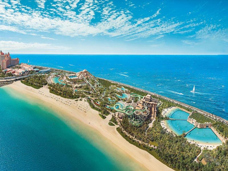 Langer Sandstrand mit blauem Meer, Hotelanlagen, Pools und bewaldeten Bereichen unter wolkigem Himmel.