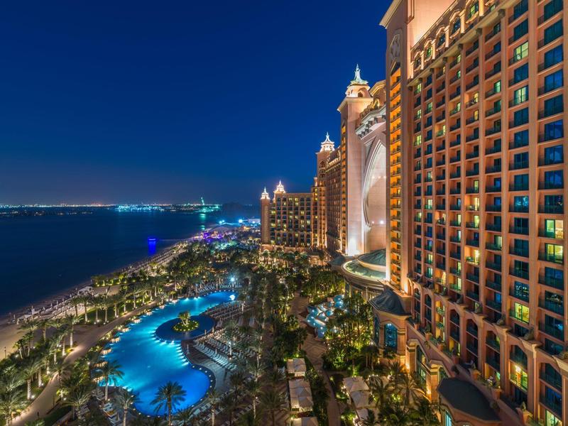 Nächtliche Stadtansicht mit beleuchtetem Pool, Hochhäusern und Meeresufer unter klarem Himmel.