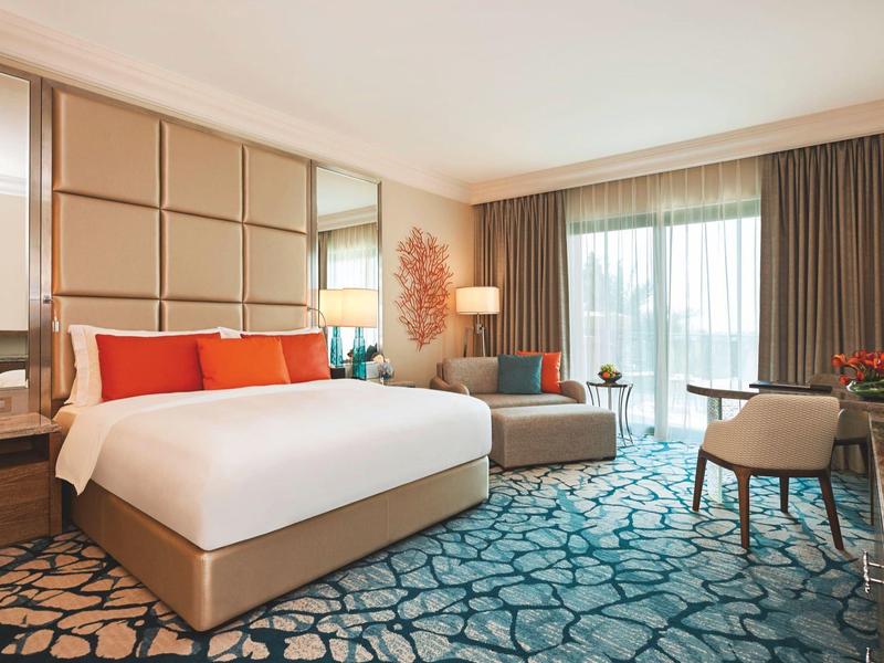 Helles Hotelzimmer mit großem Bett, Teppich mit Muster, Sessel, Vorhängen und großem Fenster.