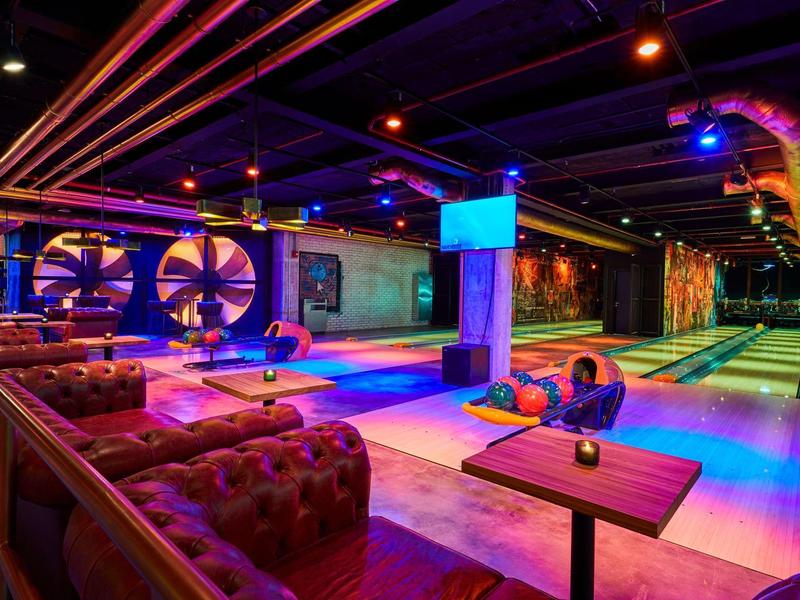 Moderne Bowlingbahn mit bunten Lichtern, Ledersofas und Tischen in entspannter Atmosphäre.