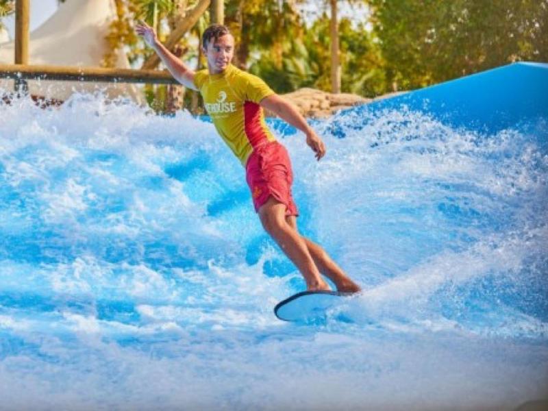 Ein Junge surft auf einer künstlichen Welle mit blauem Wasser und trägt gelbes Shirt und rote Shorts.