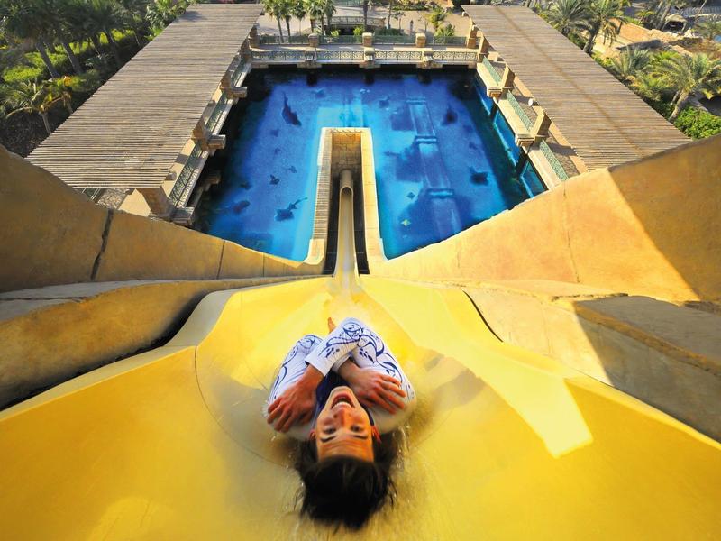 Junge rutscht auf gelber Wasserrutsche, darunter blauer Pool mit Wassertieren und Holzterrasse.