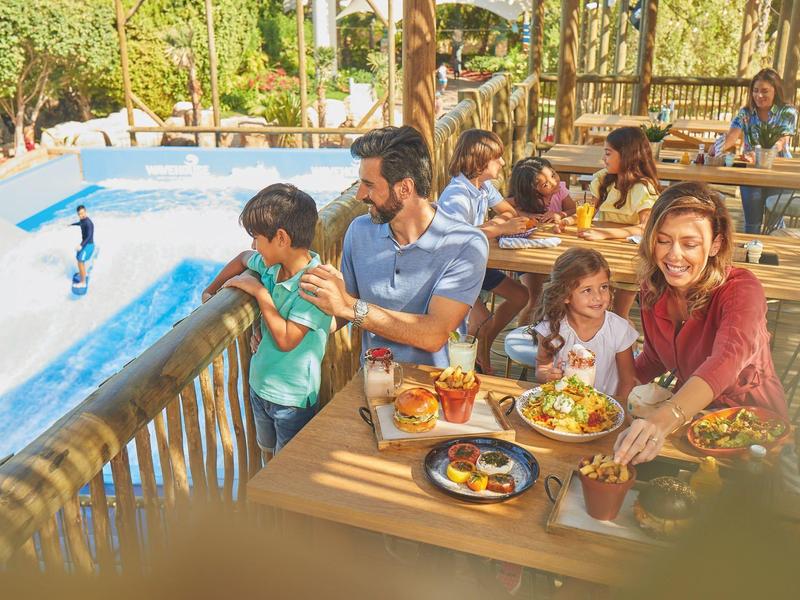 Familie mit Eltern und Kindern sitzt fröhlich am Holztisch im Außenbereich eines Restaurants.