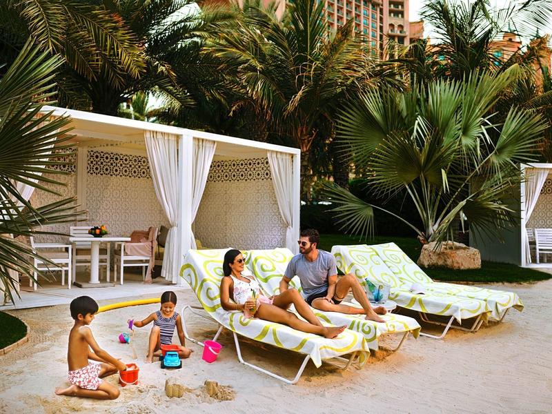 Ein sonniger Strand mit Liegestühlen, spielenden Kindern und einem Paar unter weißen Pavillons.