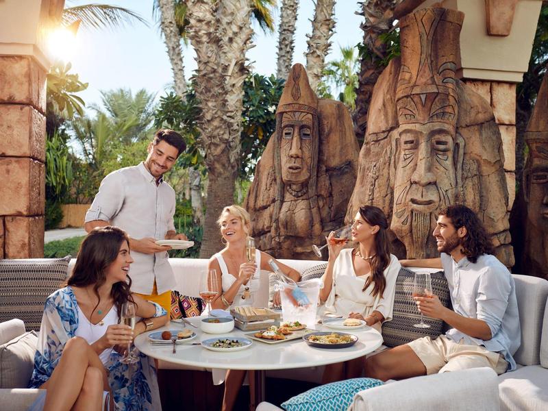 Fünf Menschen genießen Essen und Getränke an einem runden Tisch im Außenbereich mit Tropendekor.