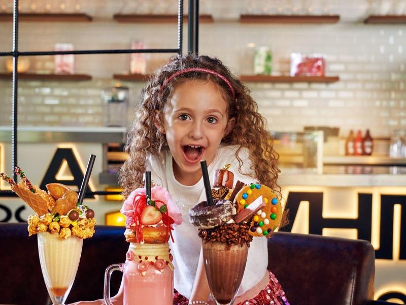 Ein fröhliches Mädchen sitzt an einem runden Holztisch mit drei bunten Milchspeisen in einem Café.