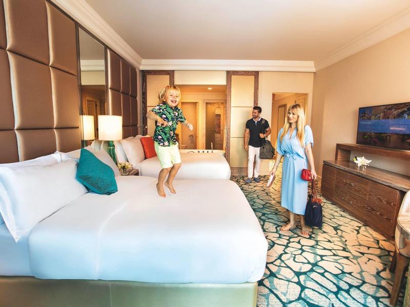 Hotelzimmer mit großem Bett, Teppichboden, Fernseher und drei Personen, zwei mit Koffern.