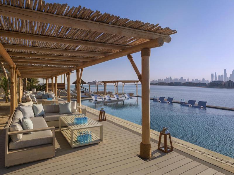 Moderner Poolbereich mit Holzpergola, bequemen Loungesesseln und Blick auf Wasser und Skyline.