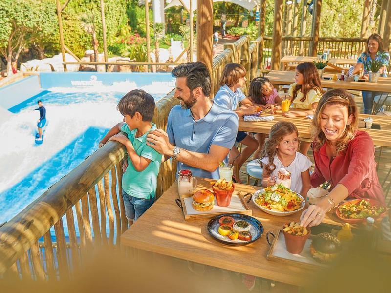 Familie sitzt draußen am Holztisch, genießt Essen und unterhält sich neben blauem Pool.