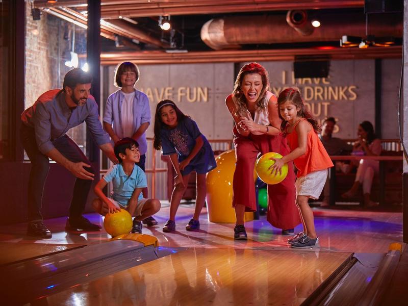 Sechs Personen bowlen in fröhlicher Runde, farbige Kugeln und warme Beleuchtung im Raum.